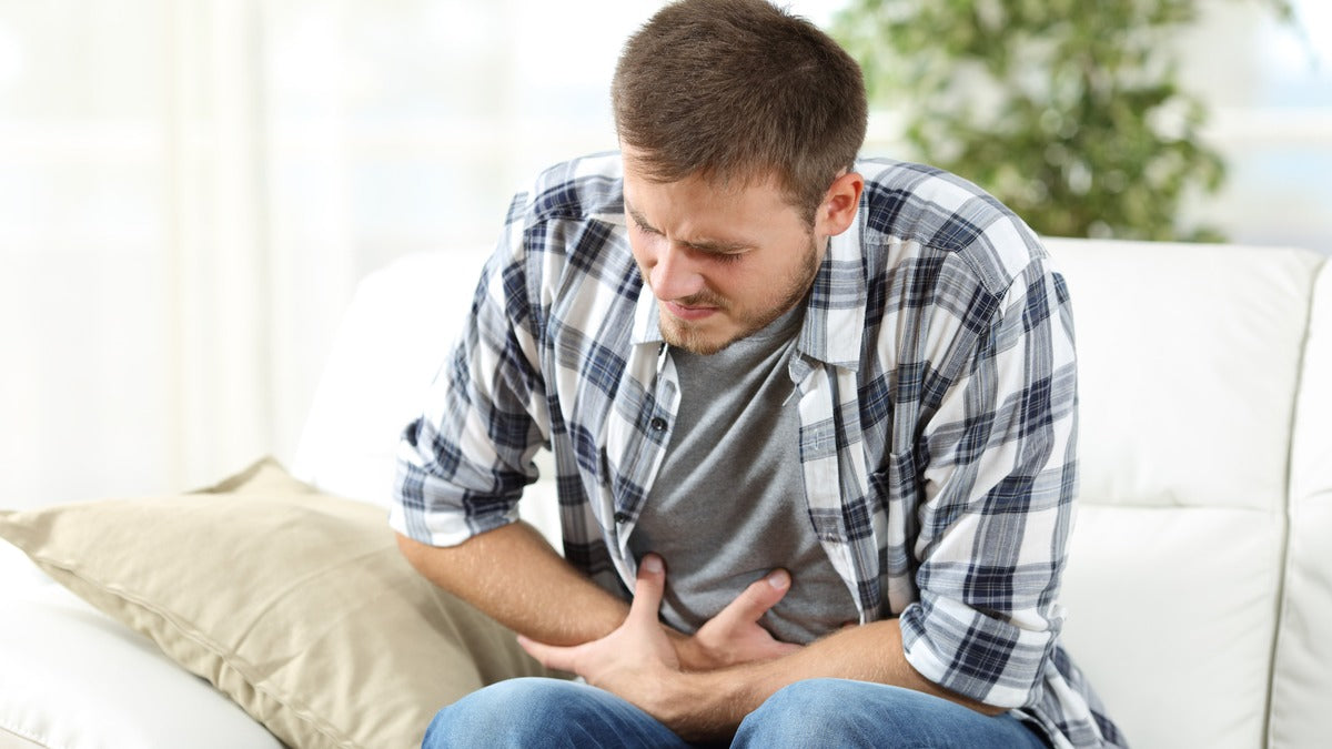 A man doubled over on his couch at home with both hands on his stomach, an hour or two after eating.